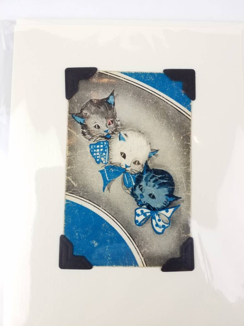 Puede incluir: Impresi&oacute;n vintage con tres gatitos de dibujos animados con lazos y detalles azules. Los gatitos tienen pelaje gris, blanco y azul. La impresi&oacute;n est&aacute; sobre un fondo gris y azul y se sujeta con esquinas negras.