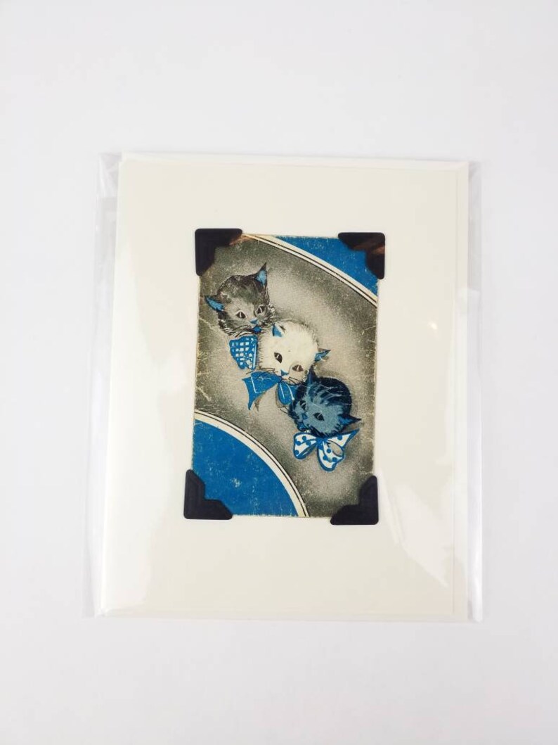 Puede incluir: Impresi&oacute;n vintage con tres caras de gatitos con lazos azules, sobre un fondo azul y gris. La impresi&oacute;n est&aacute; montada en una tarjeta blanca y asegurada con esquinas fotogr&aacute;ficas negras, en una funda de pl&aacute;stico transparente.