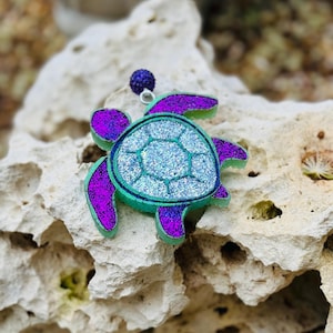 May include: A teal and purple glitter turtle charm with a silver loop for hanging. The turtle has a sparkly finish and is a cute and whimsical accessory.
