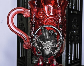 May include: A red glass goblet with a black heart and chain design. The goblet has a handle and a pointed base.