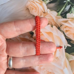 May include: Close-up of a bright red woven bracelet. The bracelet is made of a tightly woven cord, with a simple, minimalist design. The background is a soft, blurred floral pattern.