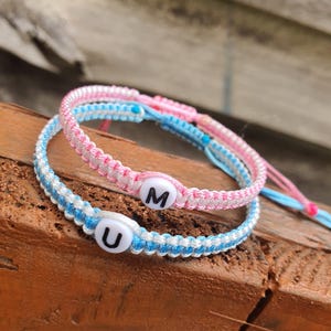 May include: Two handmade friendship bracelets. One is light blue and white, the other is pink and white. Each bracelet has a white bead with a letter on it. One says "U" and the other says "M".