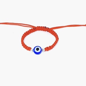 May include: Red string bracelet with a blue evil eye charm. The bracelet is adjustable and has a silver bead on either side of the charm.
