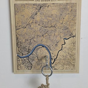 May include: Wooden wall art featuring a detailed map of Cincinnati, Ohio, with the city name and "The Queen City" text. A blue river runs through the map. A key holder with keys hangs below.