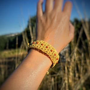Puede incluir: Una pulsera de macramé amarilla que presenta un complejo patrón de cordón tejido y pequeñas cuentas redondas de color rojo anaranjado incrustadas en el diseño. La pulsera tiene un cierre ajustable y está hecha de un material de cordón texturizado.