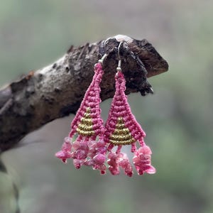 Puede incluir: Un par de pendientes triangulares de macramé con cordón rosa, detalles en hilo dorado metálico y fragmentos de piedras preciosas rosas colgantes. Los pendientes cuelgan de ganchos de metal plateado con nudos detallados.