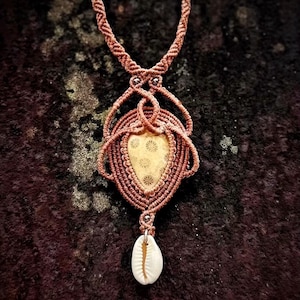 Puede incluir: Un collar de macramé con un colgante en forma de lágrima que presenta una piedra beige con patrón de estrellas, rodeada por un intrincado tejido de cordón color cobre. Una pequeña concha de cauri blanca y ovalada cuelga del colgante.