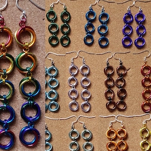 Może przedstawiać: Zestaw 10 par wiszących kolczyków wykonanych z metalowego drutu. Kolczyki są w różnych kolorach, w tym zielonym, niebieskim, srebrnym, złotym, czerwonym i brązowym. Każdy kolczyk ma trzy pierścienie.