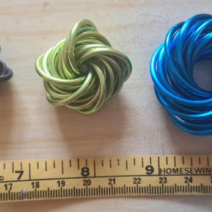 Pode incluir: Três esculturas decorativas de arame metálico em preto, verde e azul. A escultura preta tem formato de nó, a verde é um nó torcido e a azul tem um design circular. Uma régua mostra as medidas em centímetros.