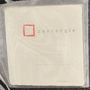 May include: Two small, rectangular packages in sheer white drawstring bags. The packages are white with the word "zentangle" and a red square printed on one. The bags are tied with white ribbons.
