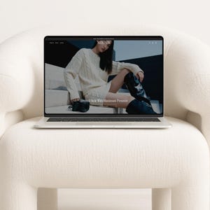 A laptop open to a fashion website homepage, resting on a textured white armchair.