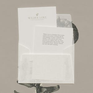 K&ouml;nnte beinhalten: Ein wei&szlig;er Umschlag mit zwei wei&szlig;en Karten. Die gr&ouml;&szlig;ere Karte zeigt den Text Wilder Lore und ein kleines Blumen-Symbol. Die kleinere Karte enth&auml;lt einen Textabsatz, der ein florales Studio beschreibt, das in der Poesie der Bl&uuml;ten verwurzelt ist.