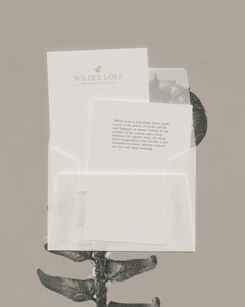 K&ouml;nnte beinhalten: Ein wei&szlig;er Umschlag mit zwei wei&szlig;en Karten. Die gr&ouml;&szlig;ere Karte zeigt den Text Wilder Lore und ein kleines Blumen-Symbol. Die kleinere Karte enth&auml;lt einen Textabsatz, der ein florales Studio beschreibt, das in der Poesie der Bl&uuml;ten verwurzelt ist.