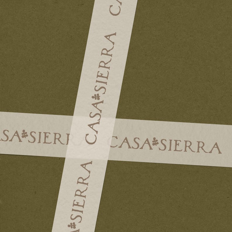 K&ouml;nnte beinhalten: Eine olivgr&uuml;ne Oberfl&auml;che, die mit zwei sich kreuzenden Streifen aus halbtransparentem wei&szlig;em Papierband umwickelt ist. Das Band zeigt den wiederholten Text CASA SIERRA in einer dunkelbraunen Serifenschrift, getrennt durch ein kleines Blumensymbol.