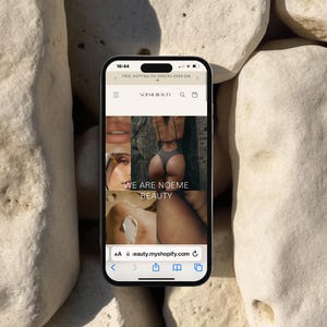 May include: A screenshot of a mobile phone displaying a website for a beauty brand called "Noeme Beauty". The website features a collage of images with the text "We are Noeme Beauty" and the website address "eauty.myshopify.com".