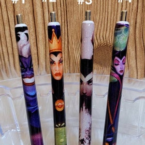 May include: Four black pens with different Disney villain characters printed on them. The pens are arranged in a row on a clear plastic stand. The characters are Ursula, Maleficent, Cinderella's stepmother, and Maleficent again. The numbers 1 through 4 are written above the pens.