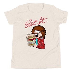 May include: Beige t-shirt with a cartoon illustration of a person holding a large sandwich. The person has brown curly hair, glasses, and a red jacket. The text "Eat It" is written above the illustration in red.