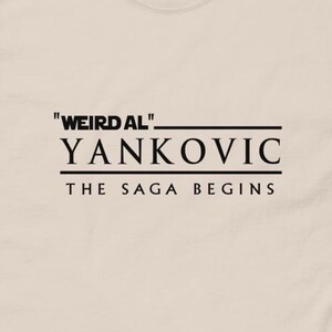 May include: A beige t-shirt with black text that reads "Weird Al" Yankovic The Saga Begins.