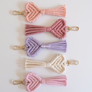 以下が含まれることがあります： ピンク、紫、クリーム色のマクラメのハート型キーホルダー5つ。各キーホルダーには、金色の留め金とタッセルが付いています。キーホルダーは白い背景に垂直に配置されています。