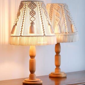 May include: Two matching table lamps with beige macrame shades and wooden bases. The shades have intricate woven patterns and tassels, casting a warm glow. The lamps are set on a wooden surface, creating a cosy ambiance.