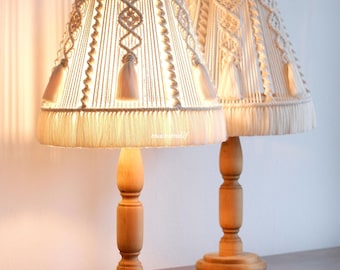 May include: Two matching table lamps with beige macrame shades and wooden bases. The shades feature intricate woven patterns and tassels, casting a warm glow. The lamps are set on a wooden surface, creating a cozy ambiance.