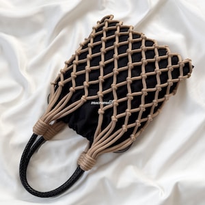 Può includere: Una borsa in tessuto nero con un rivestimento esterno in corda macramè beige in un motivo a nodi a diamante. La borsa include due manici intrecciati neri fissati alla struttura in corda. Il testo macrameli è stampato in bianco sul tessuto.