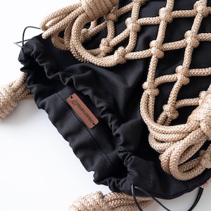 May include: Black drawstring bag with a beige rope net overlay. The bag features a leather handle and a small brown leather tag with text. The rope is knotted, creating a decorative pattern. The bag is on a white background.