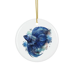 May include: A round, white ceramic ornament featuring a watercolor painting of a blue betta fish. The fish has flowing fins and is set against a backdrop of blue and purple watercolor splashes. A gold string is attached for hanging.