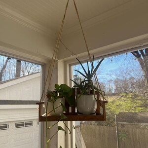 May include: A hanging wooden plant shelf with two white ceramic pots, each holding a green plant. The shelf is suspended by a natural rope from the ceiling. The plants are positioned in front of a window.