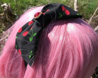 May include: A black headband with a cherry print and a red rhinestone bow. The headband is worn on a pink wig.