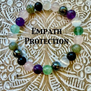 May include: A beaded bracelet with various colored gemstones, including green, purple, pink, and black, arranged in a circular pattern. The text "EMPATH PROTECTION" is displayed in the center. The bracelet is set against a carved wooden background.