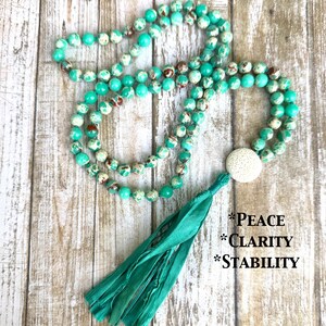 May include: A turquoise beaded necklace with a white stone accent and a teal tassel. The necklace is displayed on a light wood surface. Text on the right side of the image reads: *PEACE *CLARITY *STABILITY.