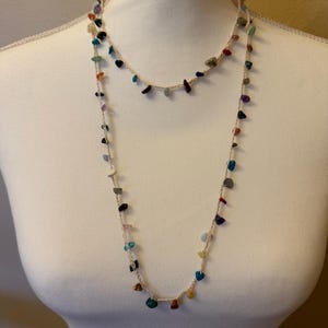 May include: A two-strand necklace featuring small, colorful gemstone beads strung on a beige cord. The gemstones are various shapes and colors, including blue, green, red, and purple. The necklace is displayed on a white mannequin.