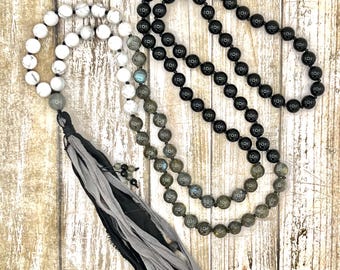 Può includere: Tre collane di perline con diverse perline di pietra e un nappa. Una collana presenta perline bianche, un'altra ha perline nere e la terza ha perline grigie e iridescenti. La nappa è nera e grigia.