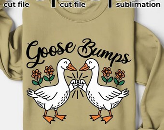 May include: A tan sweatshirt with the words "Goose Bumps" in black script. Two white cartoon geese are bumping fists, with orange beaks and feet. Orange and green flowers are on either side of the geese. The top of the image has the words "SVG | EPS | PNG" in black.