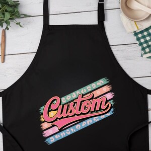 Op de afbeelding: Zwarte schort met het woord "Custom" in een retro lettertype. Het woord is roze en geel, met een achtergrond van blauwe, oranje en roze strepen. De schort heeft een nekband en strikbanden.