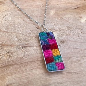 Puede incluir: Un collar plateado con un colgante rectangular. El colgante está relleno de cuadrados coloridos y texturizados en rosa, amarillo, azul y rojo. El collar se muestra sobre una superficie de madera.