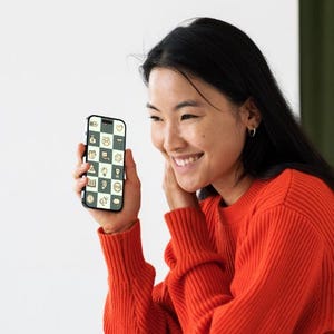 Może przedstawiać: Smartfon z czarnym etui jest trzymany przed osobą. Ekran telefonu wyświetla wz&oacute;r szachownicy z r&oacute;żnymi ikonami w kolorze białym i ż&oacute;łtym. Osoba ma na sobie jasnopomarańczowy sweter i uśmiecha się.
