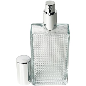May include: A clear glass perfume bottle with a silver spray top and a separate silver cap. The bottle has a rectangular shape with a textured, grid-like pattern on the surface. The cap is cylindrical.