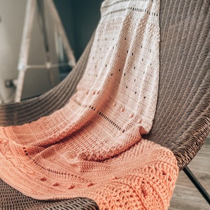 May include: A peach and cream-colored knitted blanket draped over a wicker chair. The blanket features intricate patterns and textures, with a gradient effect from cream to peach. The chair has a woven design.
