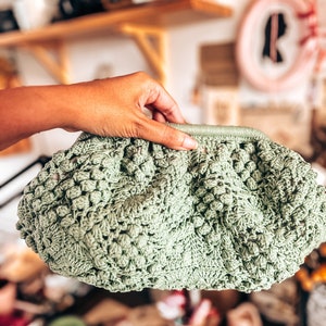 Può includere: Una borsa di uncinetto verde chiaro con un design testurizzato e gonfio. La borsa è tenuta da una mano.