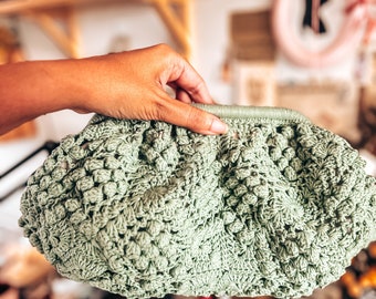 May include: A light green crocheted bag with a textured, puffy design. The bag is held by a hand.