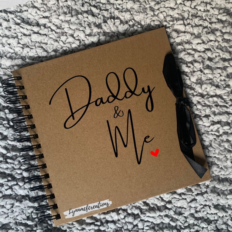 May include: A brown scrapbook with a black spiral binding and a black ribbon. The cover has the text "Daddy & Me" in black cursive with a small red heart.