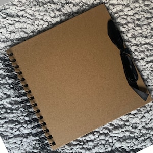 May include: A brown scrapbook with a black ribbon tied around it. The scrapbook has a spiral binding and is lying on a gray fuzzy surface.