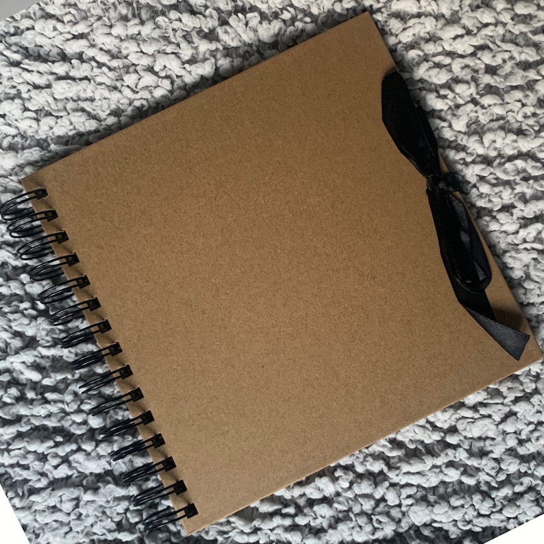May include: A brown scrapbook with a black ribbon tied around it. The scrapbook has a spiral binding and is lying on a gray fuzzy surface.