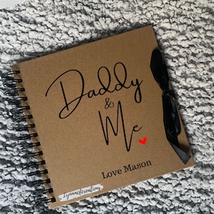 May include: A brown scrapbook with a black spiral binding and a black ribbon. The cover has the text "Daddy & Me" in black cursive with a small red heart. The text "Love Mason" is printed below the cursive text.
