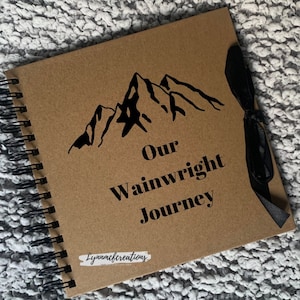 May include: A brown scrapbook with a black ribbon and a black mountain silhouette design. The text "Our Wainwright Journey" is printed on the cover.