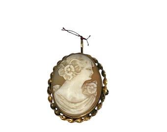 May include: Oval cameo pendant featuring a carved profile of a woman with flowers in her hair and on her dress. The cameo is set in a gold-toned, twisted rope-style frame. A small loop and string are attached at the top.