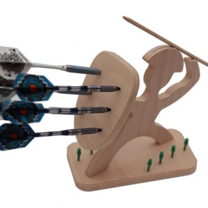 May include: Wooden dart holder shaped like a warrior holding a spear and shield. The warrior is standing on a base with small green pegs. Four darts are stuck into the shield.