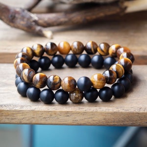 May include: Two beaded bracelets. One bracelet features tiger's eye beads, and the other has black onyx beads. The bracelets are displayed on a wooden surface. The tiger's eye beads have a mix of brown and gold tones.
